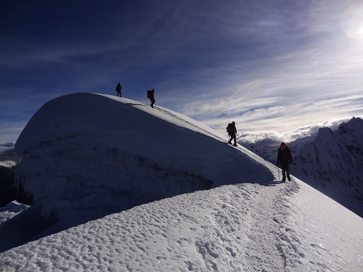 Four mountaineers are silhouetted against a twilight sky on a snowy summit