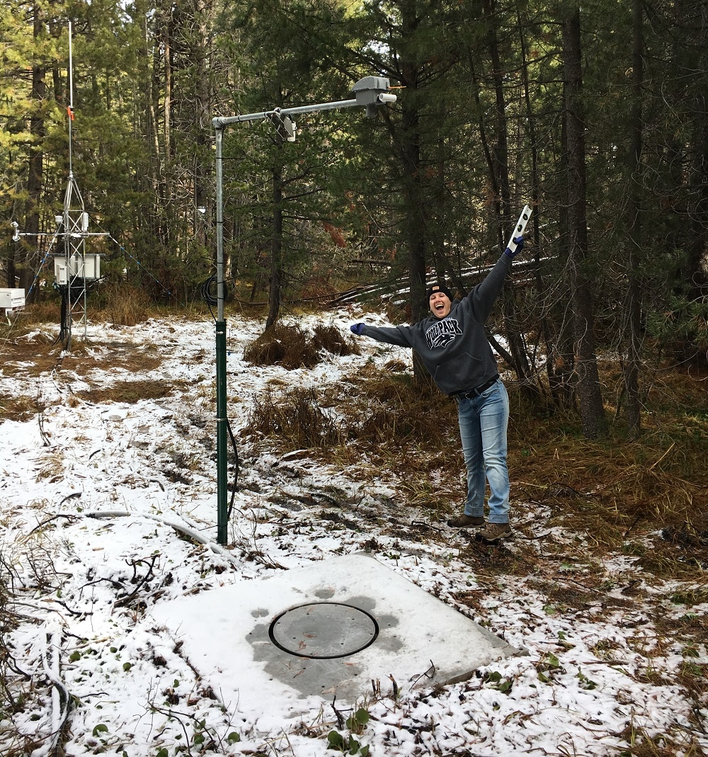 A UNR student poses with a big grin arms outstretched to the sky near a sensor device installed in snowy wooded area.