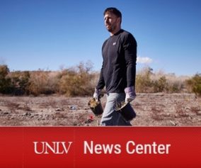 Dr. Scott Abella strides through a desert landscape. He holds potted desert vegetation in each hand.
